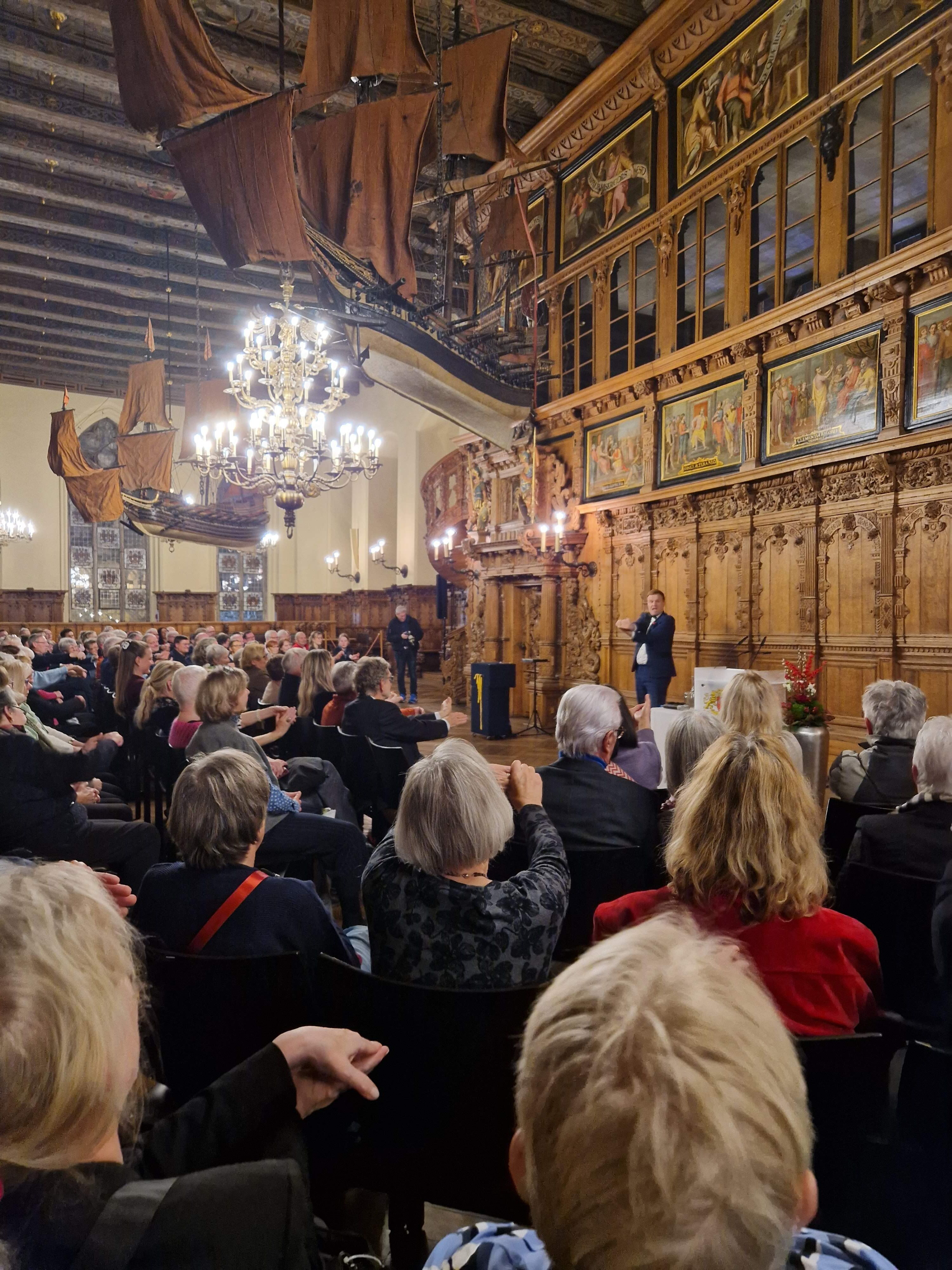 Viele Menschen sitzen in der Oberen Rathaushalle in Bremen, vorne steht ein Mann.