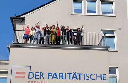 Gruppe von ca. 15 winkenden Personen auf einem Balkon eines Gebäudes, vorne Logo der Paritätische Bremen