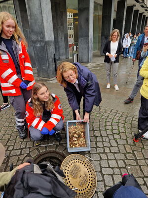 Mehrere Personen stehen um einen geöffneten Gullideckel, eine Frau hält eine Kiste mit Münzgeld in die Kamera