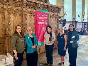5 Frauen (in der Mitte die Senatorin) im Bremer Rathaus, im Hintergrund ein pinker Aufsteller "Nachbarschaftshilfe ist mehr"
