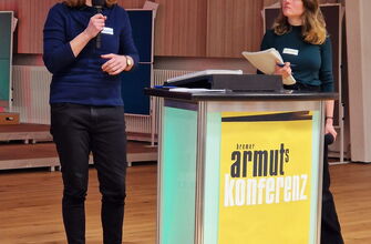 2 Frauen, eine mit Mikro an einem Rednerpult mit Plakat Armutskonferenz