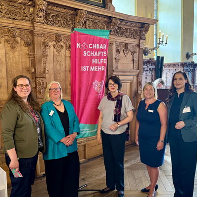 5 Frauen (in der Mitte die Senatorin) im Bremer Rathaus, im Hintergrund ein pinker Aufsteller "Nachbarschaftshilfe ist mehr"