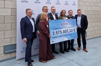 7 Personen in Anzügen halten einen großen Scheck über 2,757 Mio. Euro in der Hand