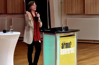 Frau mit Mikro an einem Rednerpult mit Plakat Armutskonferenz