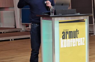 Mann mit Mikro an einem Rednerpult mit Plakat Armutskonferenz