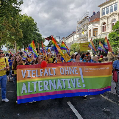 großes buntes Banner "Vielfalt ohne Alternative" und Logo des Paritätischen, dahinter viele Menschen mit bunten Flaggen bei einer Demo (CSD)