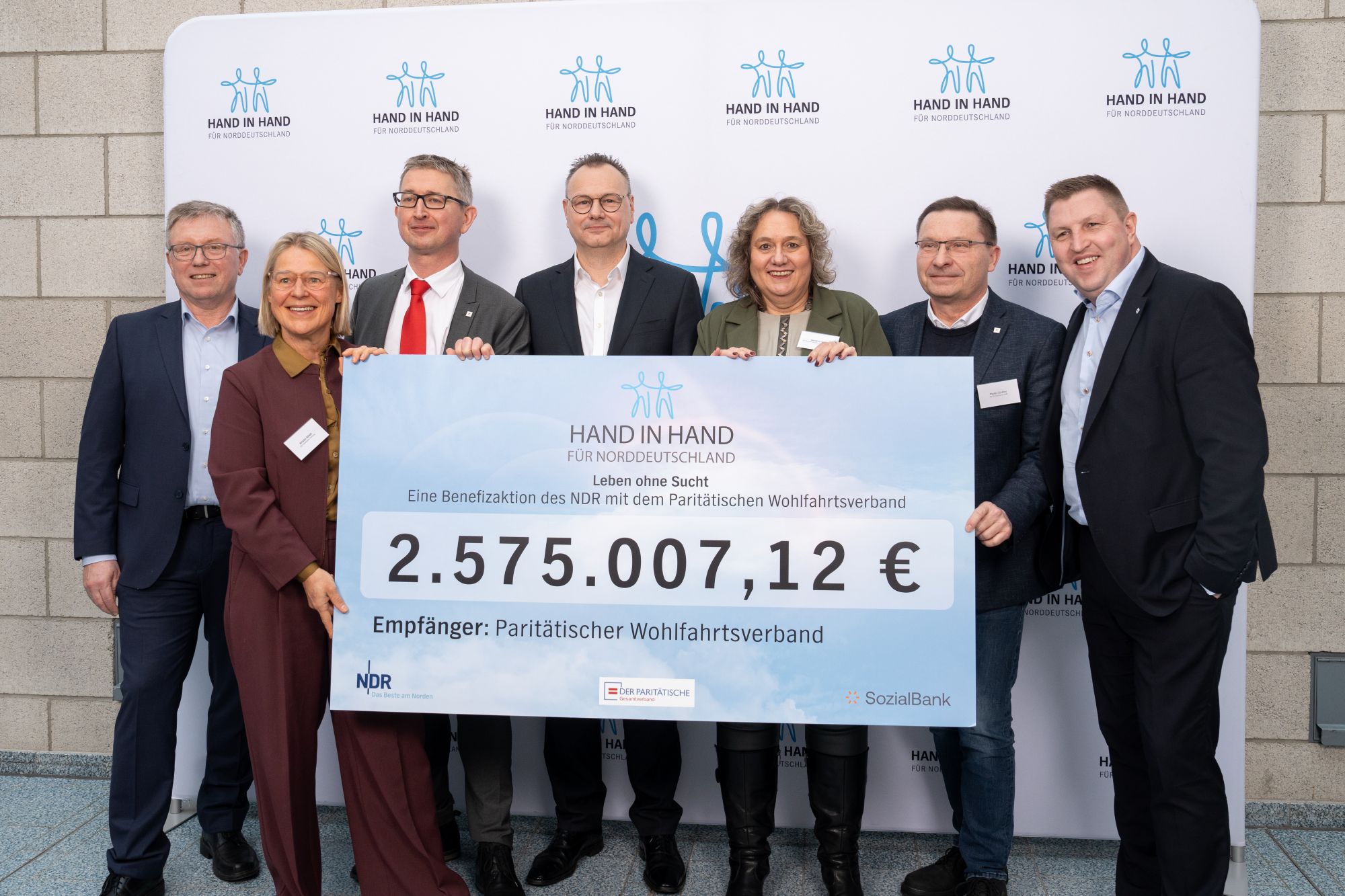 7 Personen in Anzügen halten einen großen Scheck über 2,757 Mio. Euro in der Hand
